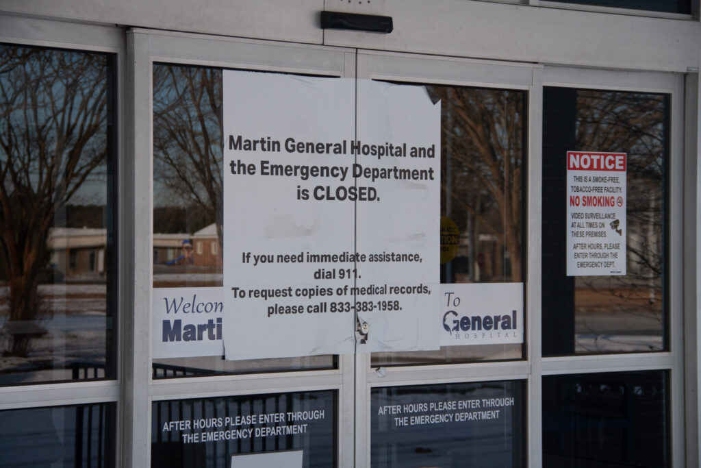 A white sign with bold black lettering is stuck to the doors of the now-closed Martin General Hospital. The sign reads "Martin General Hospital and the Emergency Department is CLOSED. If you need immediate assistance dial 911." It also instructs residents to call a number to request copies of medical records.