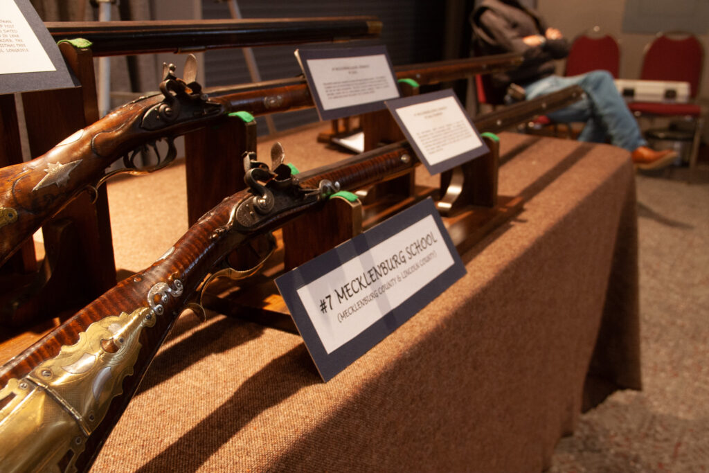 An antique longrifle from the Mecklenburg school sits prominently on a display table. The gun is made of a dark striped wood with metal detailing at the end.