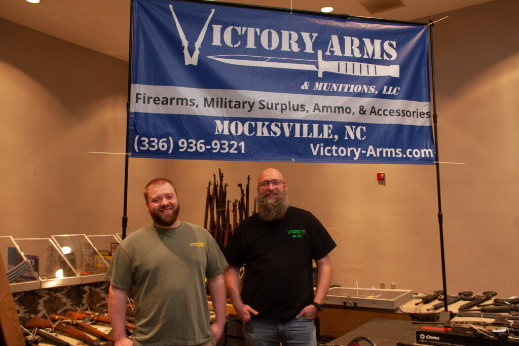 Andrew Austin stands in front of the large blue sign advertising his gun and ammo store alongside his fellow vendor.