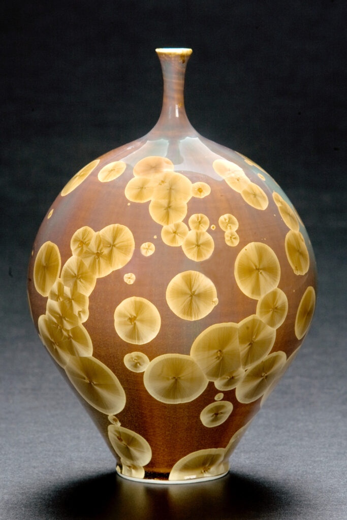 A brown pot with a wide base and a skinny neck. The pot is sporadically covered in yellow crystalline cyrcles of varying sizes.