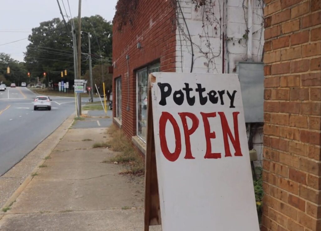 A white A-frame sign is on a sidewalk with a handpainted message of "Pottery OPEN" in large letters. The sidewalk walks by brick buildings and a nearly empty road.