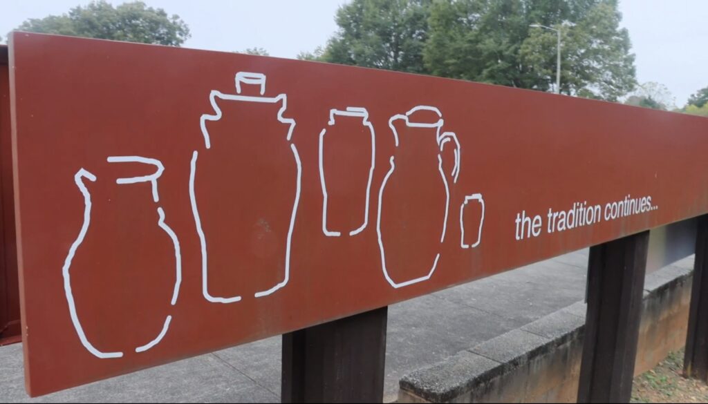 A long red wooden sign is engraved with a white design of five clay pots of different sizes and "the tradition continues..." in small text.