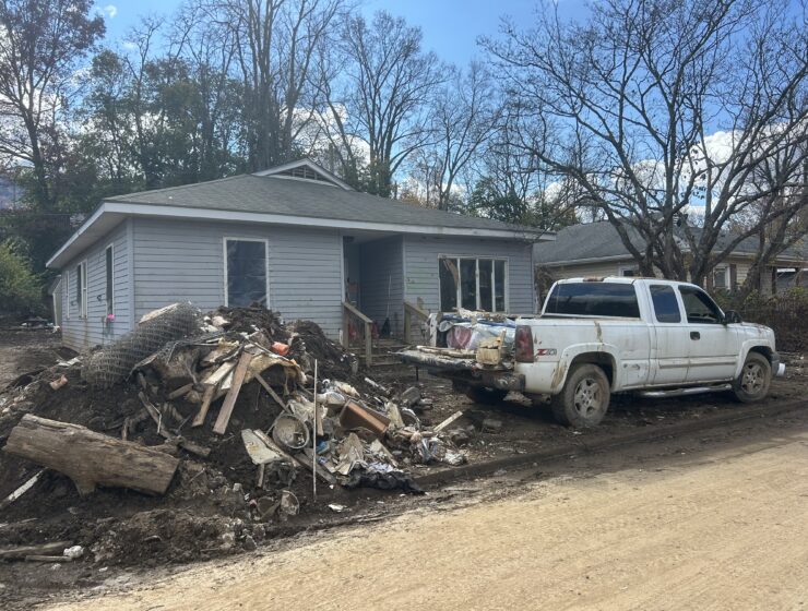 A gray one-story house in Swannanoa, N.C. A mud-covered white truck and pile of wood and rubble sit in front next to a dirt road.