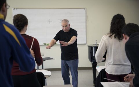 Evan Feldman stands at the front of his conducting class wearing a back T-shirt and blue pants, instructing students with his arms out.