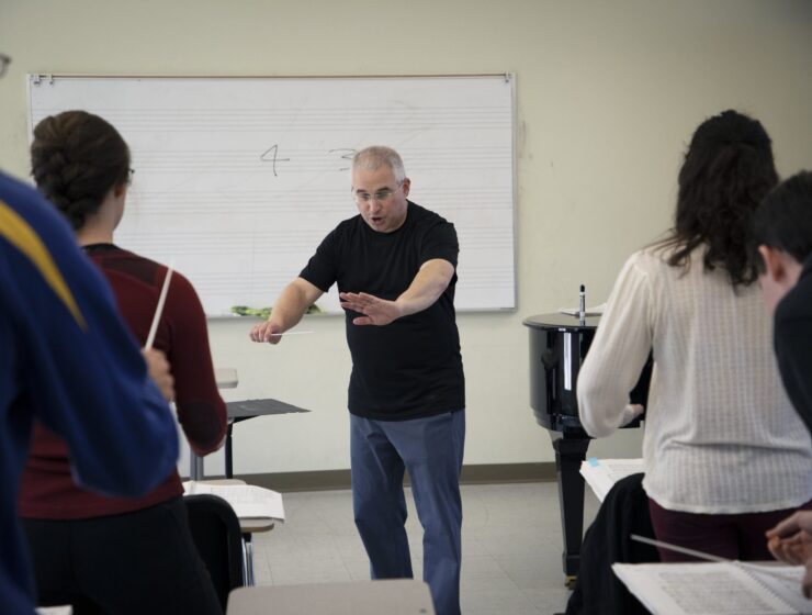 Evan Feldman stands at the front of his conducting class wearing a back T-shirt and blue pants, instructing students with his arms out.