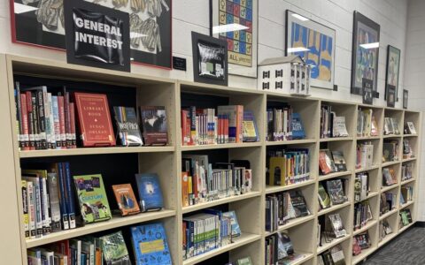 Bookshelves at Riverside High School's media center are lined with colorful books on various topics, including general interest and language and linguistics. A black sign above the shelves marks the respective books' subject area; behind it are multiple framed posters and artwork.