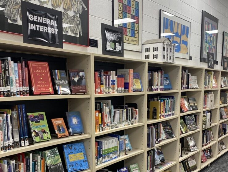 Bookshelves at Riverside High School's media center are lined with colorful books on various topics, including general interest and language and linguistics. A black sign above the shelves marks the respective books' subject area; behind it are multiple framed posters and artwork.