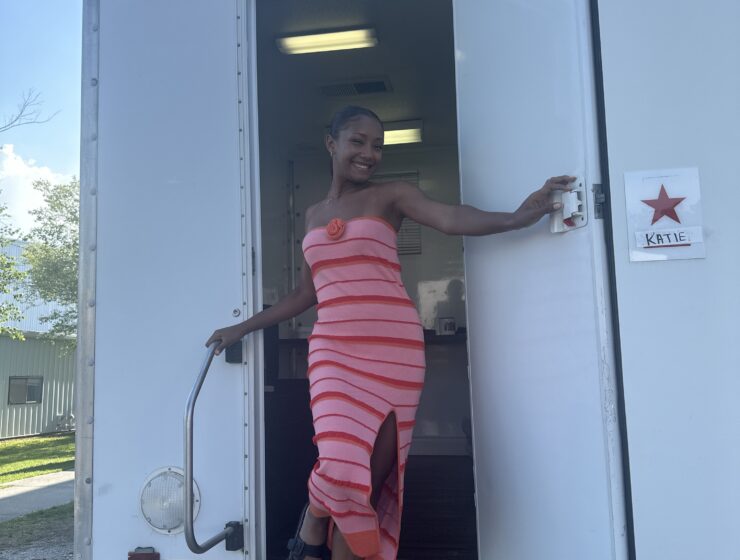 Sarah Grace Elliot poses in the open doorway of her trailer coming down the steps with a sign that reads Katie beside the ajar door.