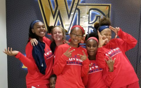 Five girls in basketball uniforms pose in front of Wake Forest logo