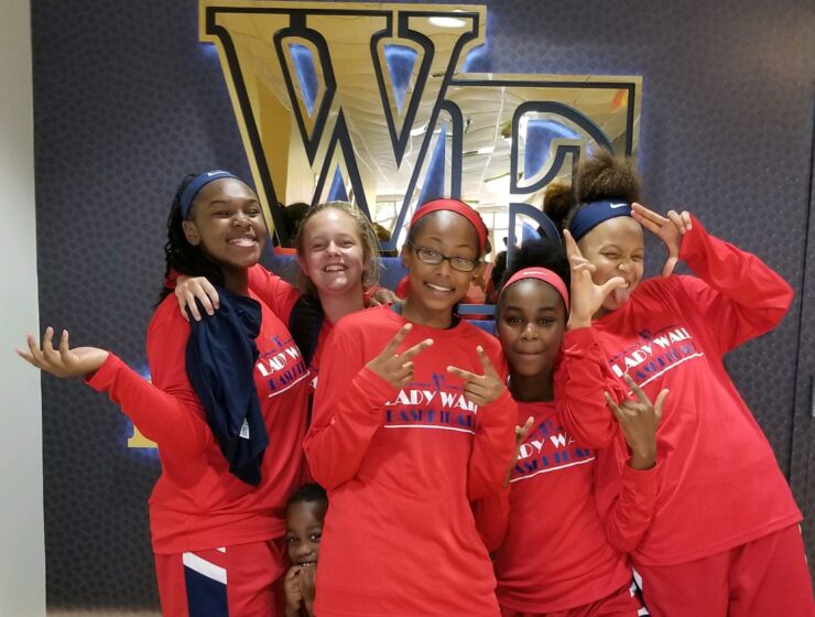 Five girls in basketball uniforms pose in front of Wake Forest logo