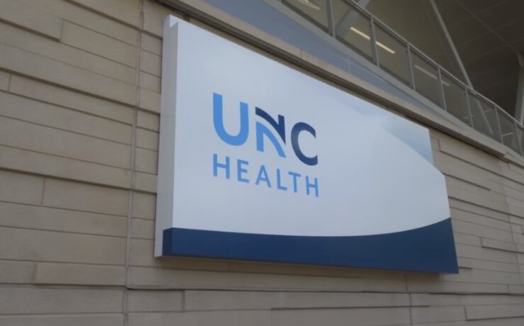 A white and blue sign on the side of a building with "UNC Health" written in light blue and dark blue letters across it.