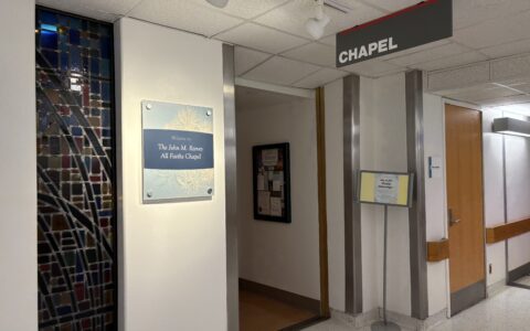 A hospital hallways has a sign hanging from the ceiling that says 'Chapel' and a sign on the hallway wall reads 'Welcome to the John M. Reeves All Faiths Chapel.