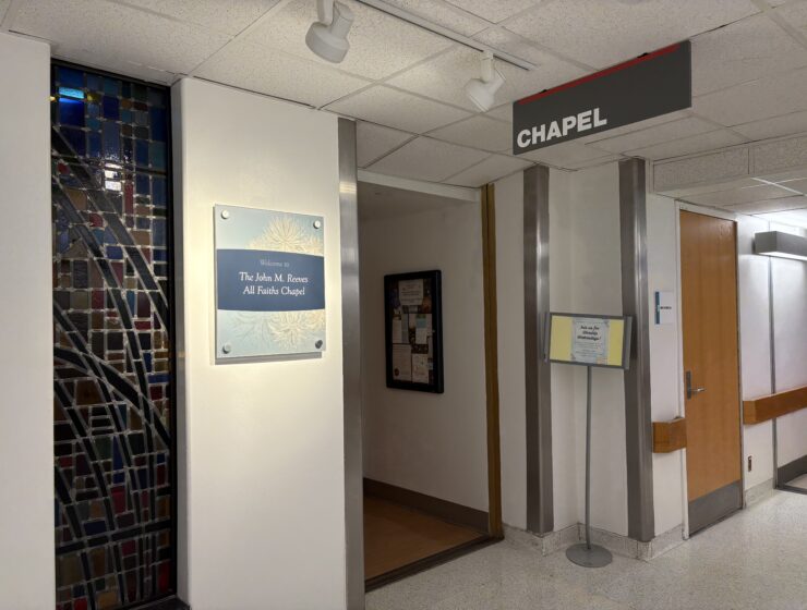 A hospital hallways has a sign hanging from the ceiling that says 'Chapel' and a sign on the hallway wall reads 'Welcome to the John M. Reeves All Faiths Chapel.