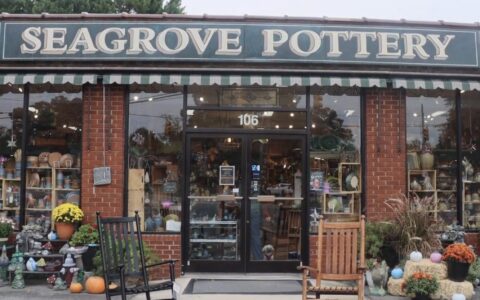 The front of a store with a sign over the top that says "Seagrove Pottery" and large windows around the exterior. Inside, there are shelves filled with pottery pieces. Two rocking chairs are positioned in front of the entrance, and arrangements of flowers and decorations are on both sides.