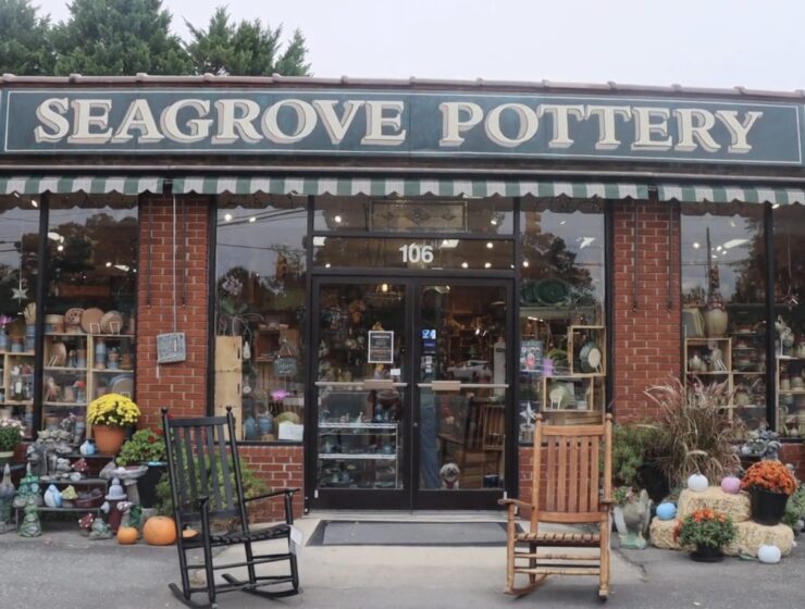 The front of a store with a sign over the top that says "Seagrove Pottery" and large windows around the exterior. Inside, there are shelves filled with pottery pieces. Two rocking chairs are positioned in front of the entrance, and arrangements of flowers and decorations are on both sides.