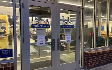 Two double glass doors of Bestway Marketplace. A paper with alternative Thanksgiving hours are taped to the doors. Through the glass walls you can see the checkout kiosks and shelves.