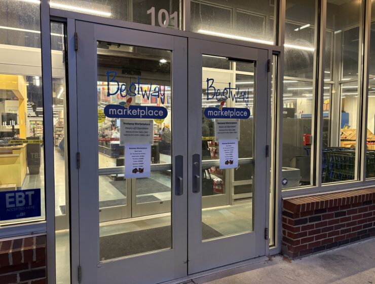Two double glass doors of Bestway Marketplace. A paper with alternative Thanksgiving hours are taped to the doors. Through the glass walls you can see the checkout kiosks and shelves.