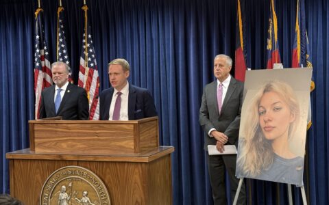 A man stands behind a podium with two men behind him on either side in front of a navy curtain. American and North Carolina state flags stand in front of the curtain, and a picture of a blonde woman is on an easel next to the podium.