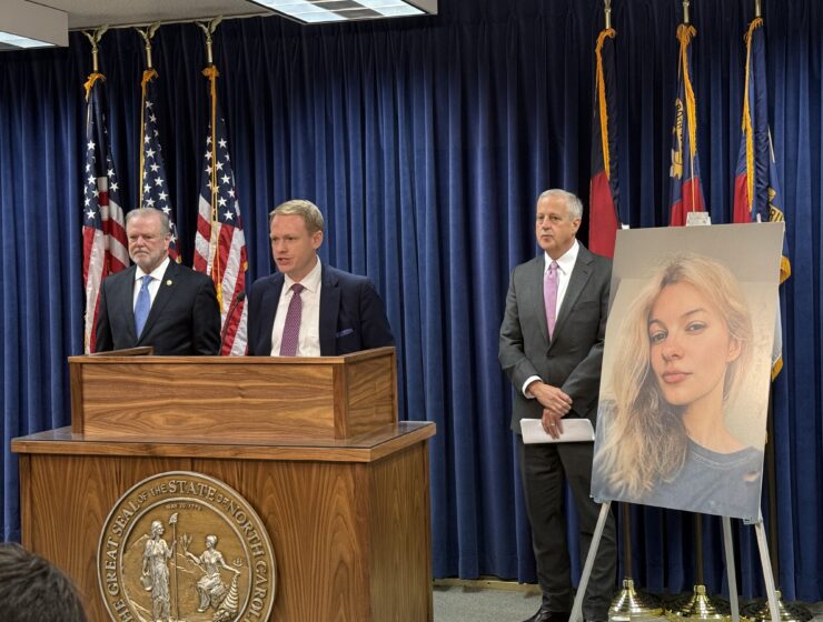 A man stands behind a podium with two men behind him on either side in front of a navy curtain. American and North Carolina state flags stand in front of the curtain, and a picture of a blonde woman is on an easel next to the podium.