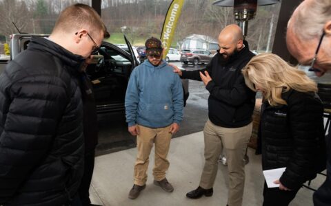A man in a black sweatshirt bows his head in prayer. One of his hands is placed on the shoulder of a man in a blue sweatshirt next to him, and his other hand is across his chest. Three others also have their heads bowed.